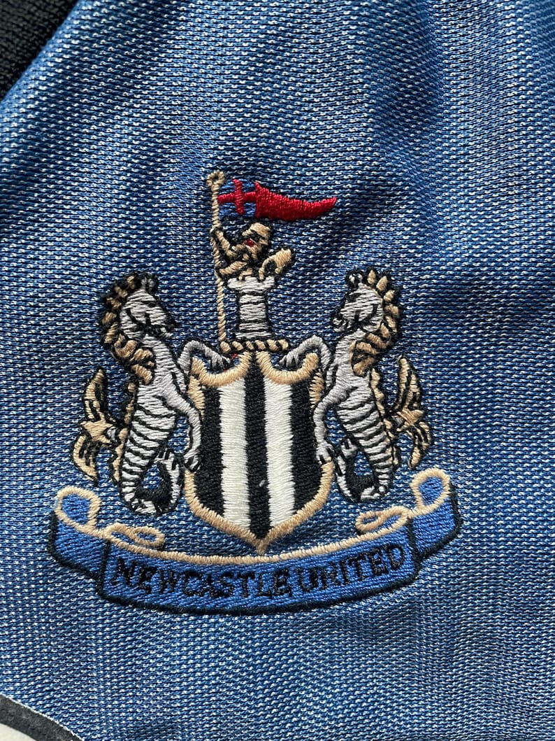 May include: Embroidered Newcastle United crest on a blue fabric. The crest features a shield with black and white stripes, flanked by two seahorses, and a banner that reads "NEWCASTLE UNITED". A red flag is at the top.