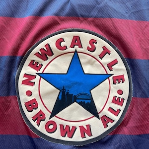 May include: A close-up of a striped jersey in burgundy and navy blue. The jersey features a circular patch with the words "NEWCASTLE BROWN ALE" around a blue star with a silhouette of a city skyline.
