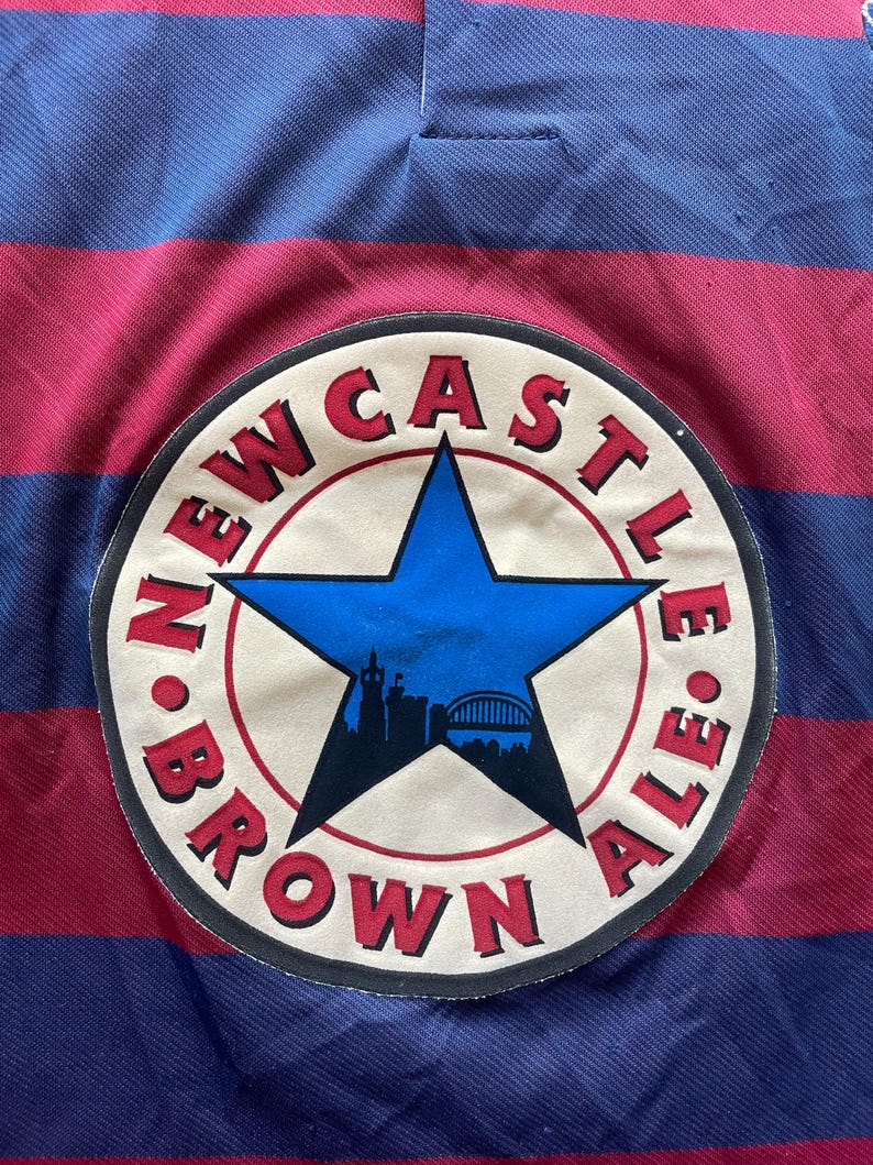 May include: A close-up of a striped jersey in burgundy and navy blue. The jersey features a circular patch with the words "NEWCASTLE BROWN ALE" around a blue star with a silhouette of a city skyline.