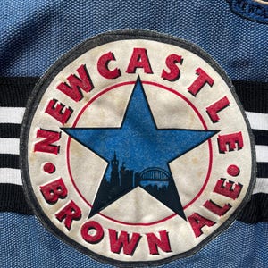 May include: Close-up of a blue and white soccer jersey featuring a circular patch. The patch has a blue star in the center with a city silhouette. The words "NEWCASTLE BROWN ALE" are in red around the star. The jersey has black and white stripes.