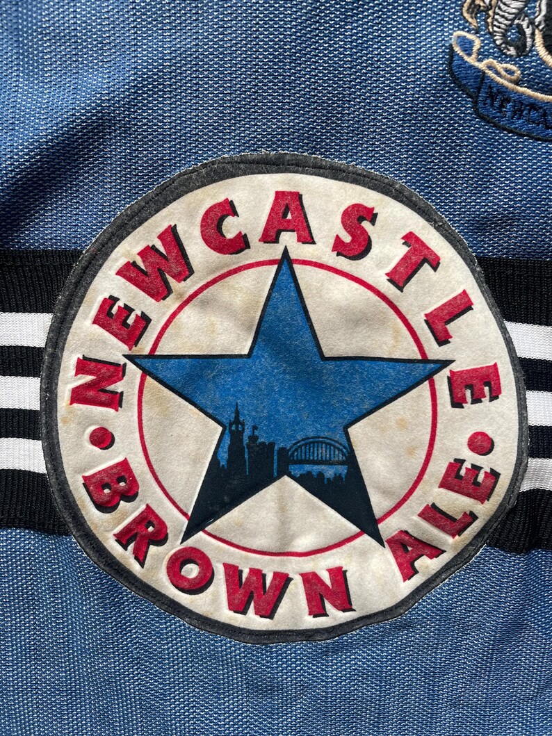 May include: Close-up of a blue and white soccer jersey featuring a circular patch. The patch has a blue star in the center with a city silhouette. The words "NEWCASTLE BROWN ALE" are in red around the star. The jersey has black and white stripes.
