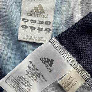May include: Close-up of two Adidas garment tags. One tag displays sizing information in multiple international standards. The other tag includes the Adidas logo, text in multiple languages, and a production date of 07/04 with the number 369698 and AHJ001.