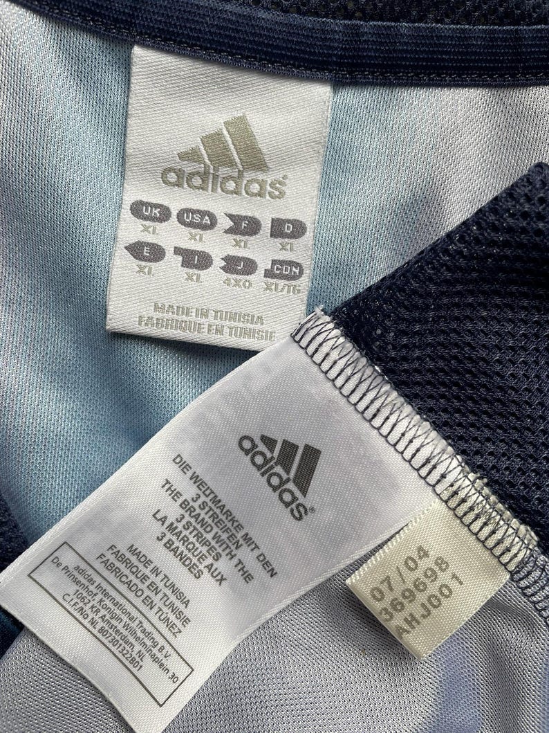 May include: Close-up of two Adidas garment tags. One tag displays sizing information in multiple international standards. The other tag includes the Adidas logo, text in multiple languages, and a production date of 07/04 with the number 369698 and AHJ001.