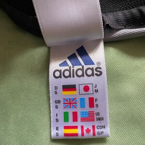 May include: Close-up of a white tag with the Adidas logo and flags representing different countries. The tag includes size indicators and the Adidas logo in blue. The background is a light green fabric.