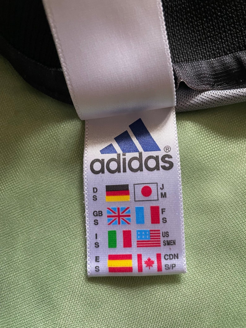 May include: Close-up of a white tag with the Adidas logo and flags representing different countries. The tag includes size indicators and the Adidas logo in blue. The background is a light green fabric.