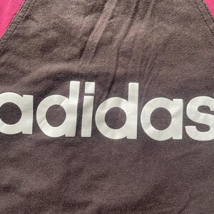 Puede incluir: Primer plano de una tela marr&oacute;n oscuro con el logotipo blanco de Adidas. La esquina superior izquierda es de color rosa intenso. El logotipo de Adidas est&aacute; centrado y ocupa la mayor parte de la imagen.