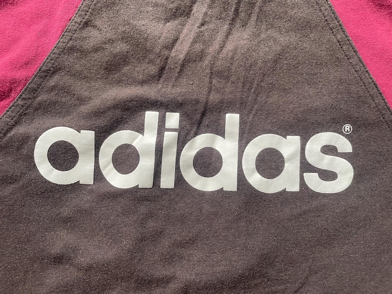 Puede incluir: Primer plano de una tela marr&oacute;n oscuro con el logotipo blanco de Adidas. La esquina superior izquierda es de color rosa intenso. El logotipo de Adidas est&aacute; centrado y ocupa la mayor parte de la imagen.