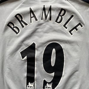 Puede incluir: Una camiseta de f&uacute;tbol gris con letras negras que dicen "BRAMBLE 19". La camiseta tiene el logotipo de la Premier League en la parte inferior.
