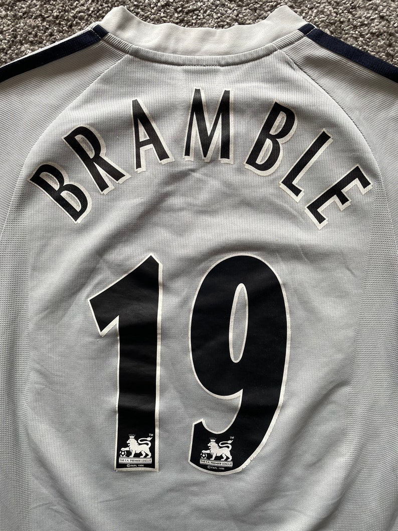 Puede incluir: Una camiseta de f&uacute;tbol gris con letras negras que dicen "BRAMBLE 19". La camiseta tiene el logotipo de la Premier League en la parte inferior.