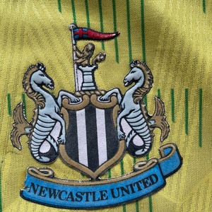 May include: A yellow and green soccer jersey with a black and white Newcastle United crest. The crest features two seahorses and a shield with black and white stripes. The text "NEWCASTLE UNITED" is on a blue ribbon.