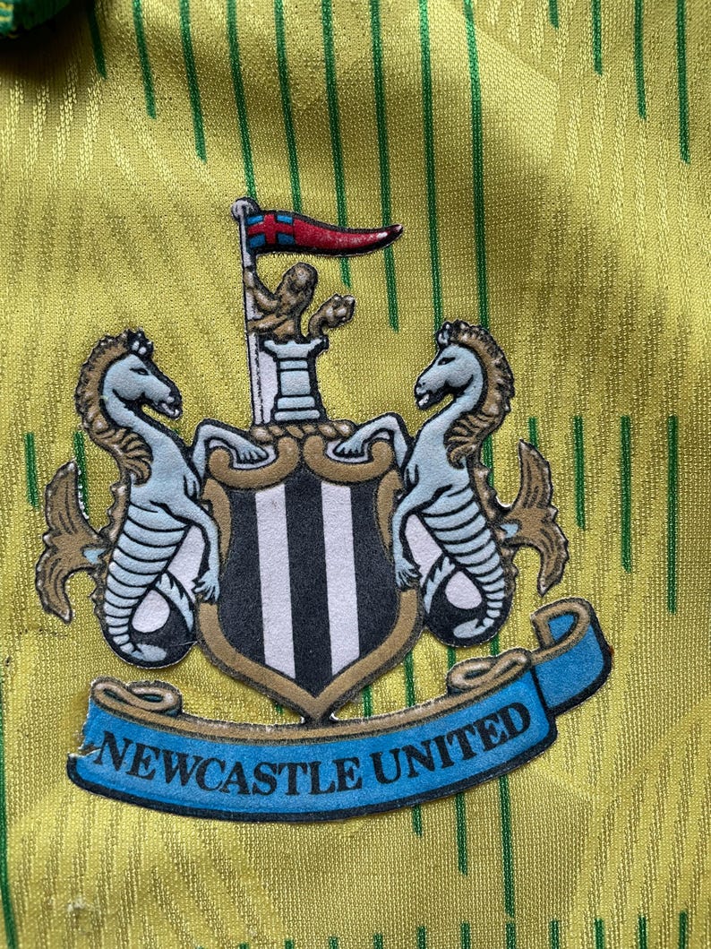 May include: A yellow and green soccer jersey with a black and white Newcastle United crest. The crest features two seahorses and a shield with black and white stripes. The text "NEWCASTLE UNITED" is on a blue ribbon.