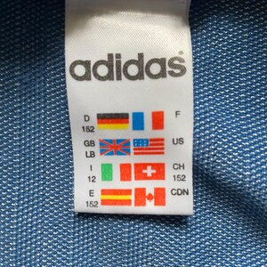 May include: A white Adidas label featuring the brand name in black. The label displays various country flags and size details. The background is a textured blue fabric.