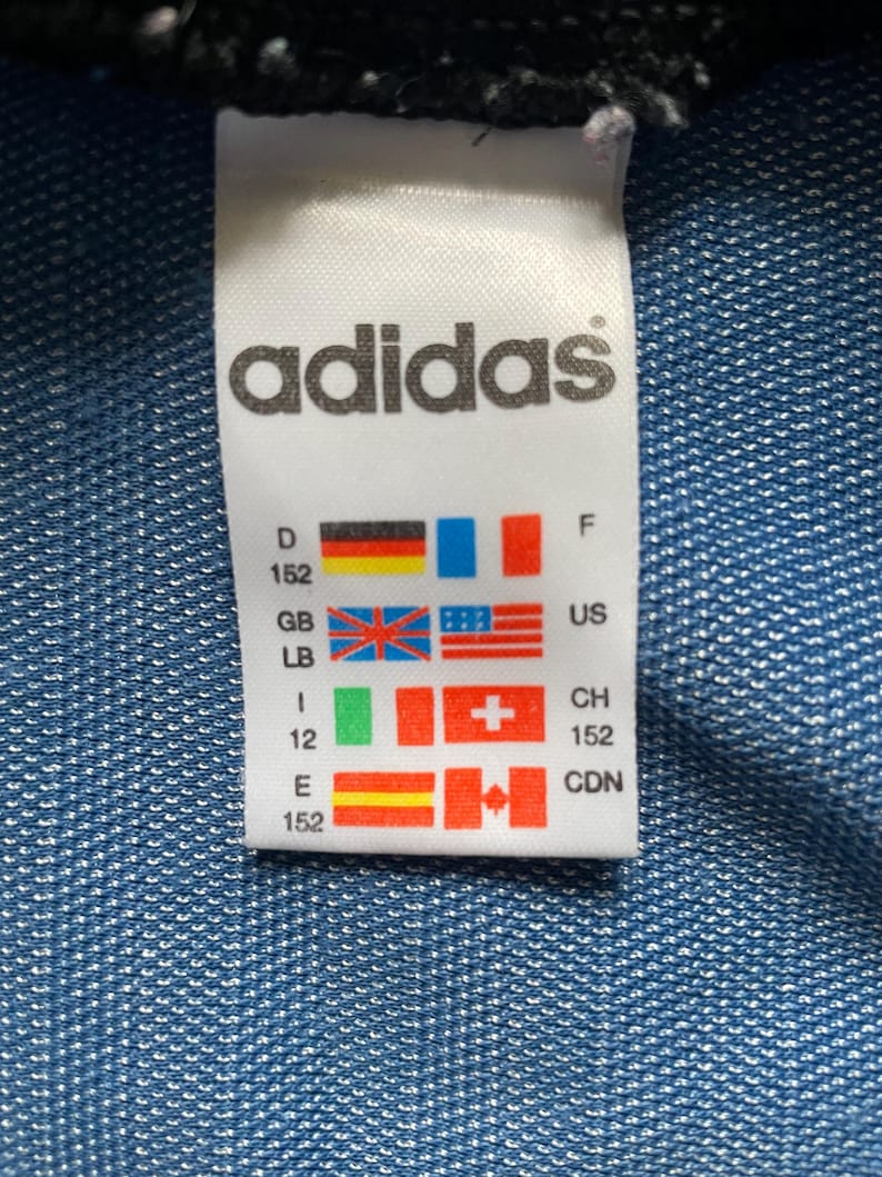 May include: A white Adidas label featuring the brand name in black. The label displays various country flags and size details. The background is a textured blue fabric.