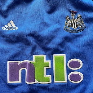 May include: A blue soccer jersey with the Adidas logo, the Newcastle United crest, and the NTL logo.