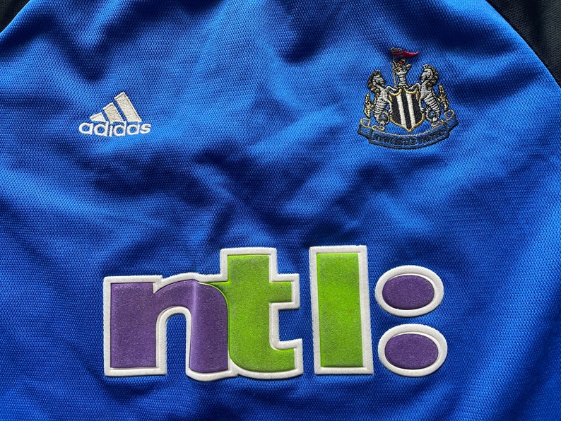 May include: A blue soccer jersey with the Adidas logo, the Newcastle United crest, and the NTL logo.