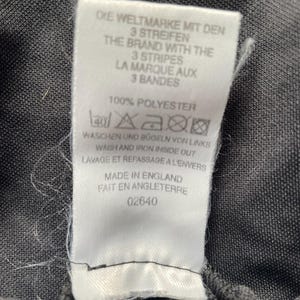 May include: A white tag with black text, including "DIE WELTMARKE MIT DEN 3 STREIFEN" and "THE BRAND WITH THE 3 STRIPES". The tag also states "100% POLYESTER" and washing instructions. The tag is attached to a dark gray fabric.