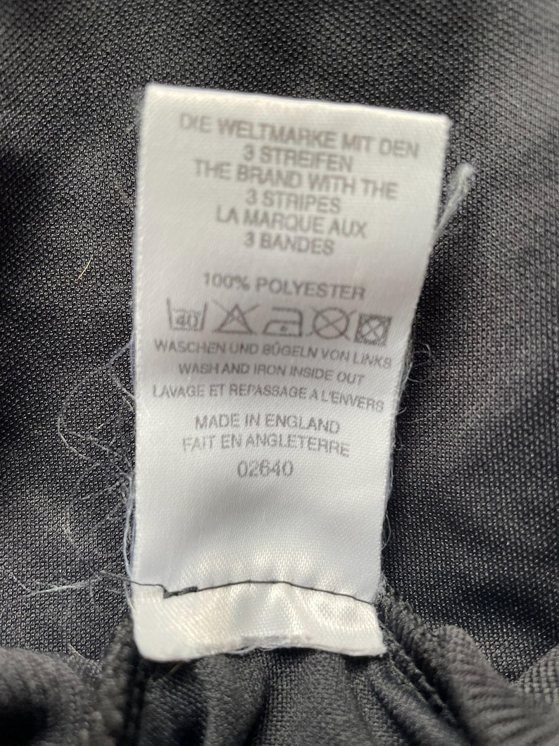 May include: A white tag with black text, including "DIE WELTMARKE MIT DEN 3 STREIFEN" and "THE BRAND WITH THE 3 STRIPES". The tag also states "100% POLYESTER" and washing instructions. The tag is attached to a dark gray fabric.