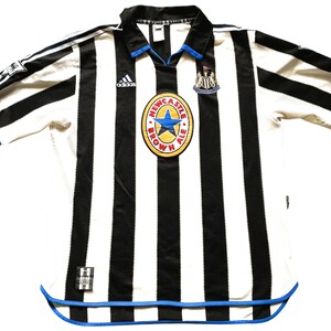 Puede incluir: Una camiseta de f&uacute;tbol a rayas blancas y negras con ribetes azules. La camiseta tiene el logotipo de Adidas en el pecho izquierdo y el logotipo de Newcastle Brown Ale en el pecho derecho. La camiseta es de talla grande.