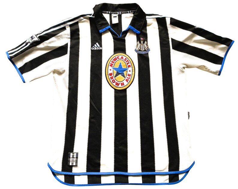 Puede incluir: Una camiseta de f&uacute;tbol a rayas blancas y negras con ribetes azules. La camiseta tiene el logotipo de Adidas en el pecho izquierdo y el logotipo de Newcastle Brown Ale en el pecho derecho. La camiseta es de talla grande.
