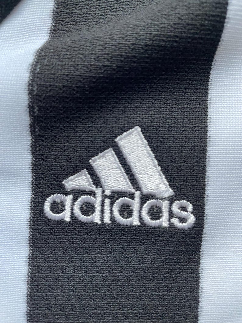 Pode incluir: Um tecido listrado branco e preto com um logotipo Adidas branco bordado em uma faixa preta.