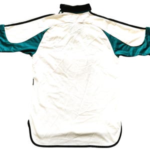 May include: White and teal athletic jersey with black trim. The jersey has a collared neckline and a curved hemline. Teal panels are on the sleeves and sides. The jersey is made of a lightweight, possibly satin-like material.