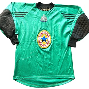 May include: A green and black long-sleeved soccer jersey with the Newcastle Brown Ale logo. The jersey has black padded sleeves and the Adidas logo. The jersey has a textured pattern and the Newcastle United crest.