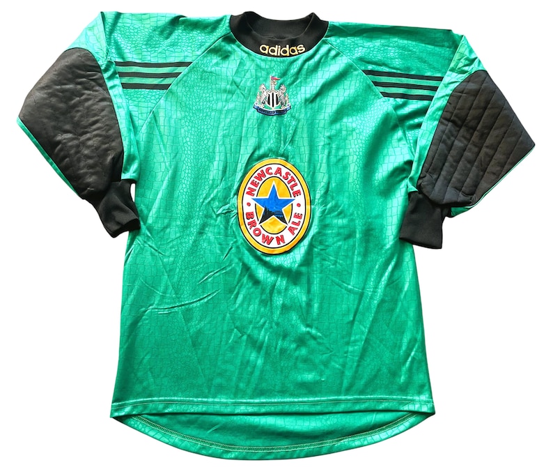 May include: A green and black long-sleeved soccer jersey with the Newcastle Brown Ale logo. The jersey has black padded sleeves and the Adidas logo. The jersey has a textured pattern and the Newcastle United crest.