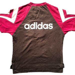 Puede incluir: Una camiseta Adidas marr&oacute;n con detalles burdeos y blancos. El logotipo de Adidas en blanco est&aacute; en el pecho. Las mangas tienen rayas blancas y paneles burdeos.