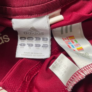 Pu&ograve; includere: Un'etichetta bianca con testo nero che recita "adidas" e informazioni sulle taglie in taglie UK, USA, F e D. L'etichetta riporta anche "Made in Tunisia, Fabrique en Tunisie".