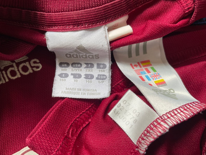 Pu&ograve; includere: Un'etichetta bianca con testo nero che recita "adidas" e informazioni sulle taglie in taglie UK, USA, F e D. L'etichetta riporta anche "Made in Tunisia, Fabrique en Tunisie".