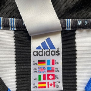 Może przedstawiać: Biała metka z czarnym tekstem, na kt&oacute;rej widnieje napis "adidas" oraz tabela rozmiar&oacute;w z r&oacute;żnymi rozmiarami krajowymi dla XXL. Metka jest przymocowana do czarno-białej, pasiastych koszulki.