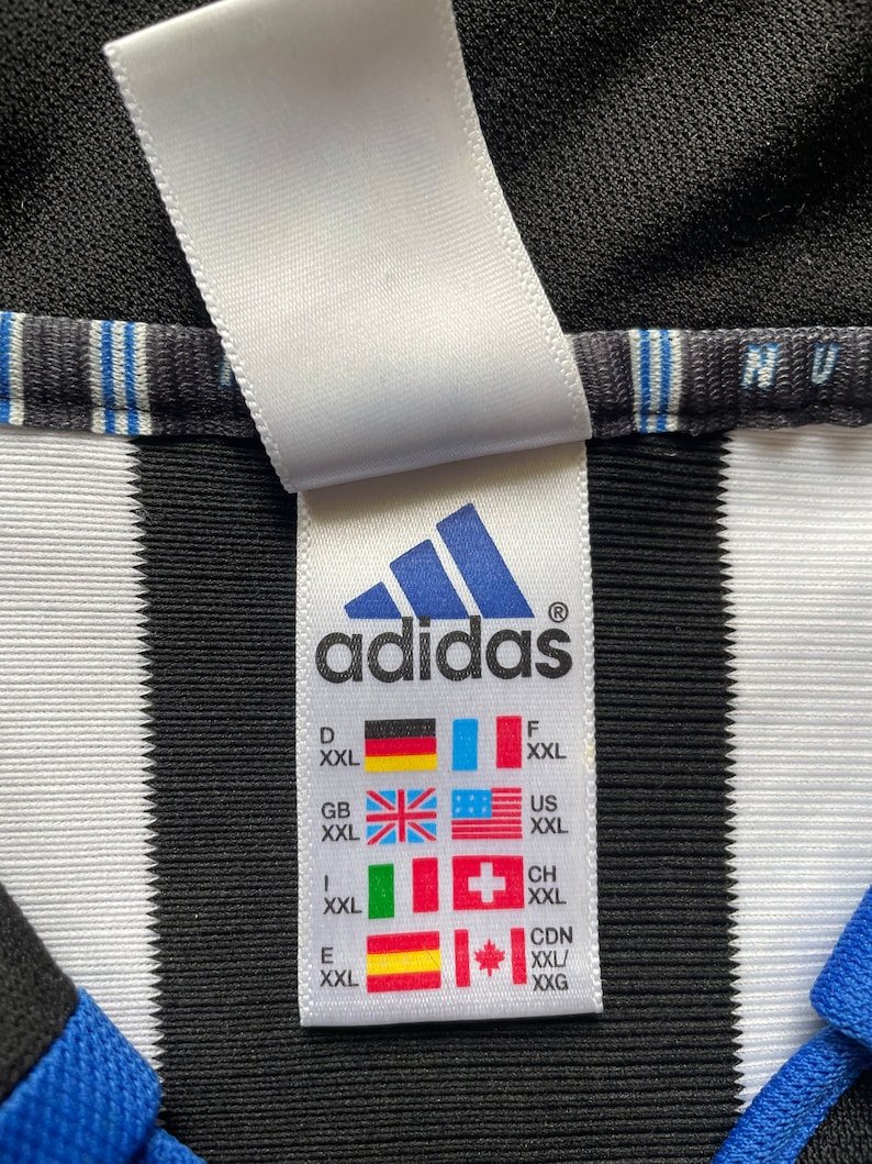 Może przedstawiać: Biała metka z czarnym tekstem, na kt&oacute;rej widnieje napis "adidas" oraz tabela rozmiar&oacute;w z r&oacute;żnymi rozmiarami krajowymi dla XXL. Metka jest przymocowana do czarno-białej, pasiastych koszulki.