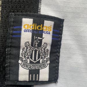 May include: Close-up of a black and white Newcastle United jersey tag. The tag features the Adidas logo in gold, the words "OFFICIAL REPLICA", and the Newcastle United crest. The jersey has black and white stripes.