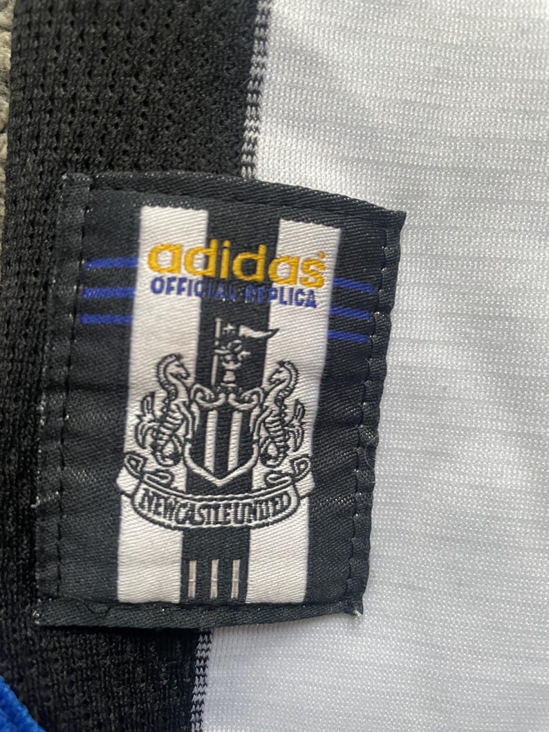May include: Close-up of a black and white Newcastle United jersey tag. The tag features the Adidas logo in gold, the words "OFFICIAL REPLICA", and the Newcastle United crest. The jersey has black and white stripes.
