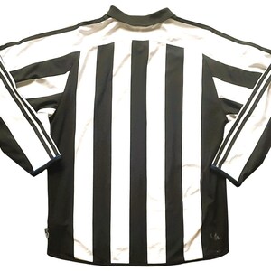 Puede incluir: Camiseta deportiva de manga larga a rayas blancas y negras. La camiseta tiene cuello y tres rayas blancas en cada manga. La parte trasera de la camiseta presenta rayas verticales blancas y negras.