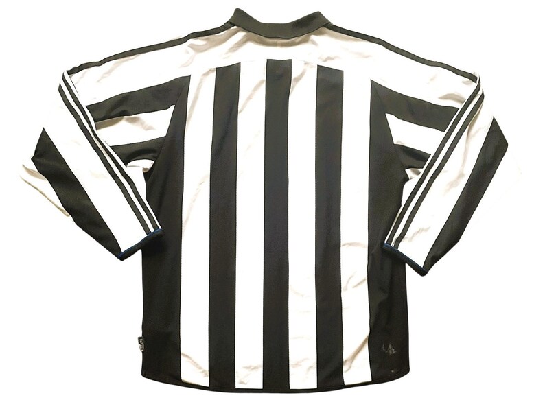 Puede incluir: Camiseta deportiva de manga larga a rayas blancas y negras. La camiseta tiene cuello y tres rayas blancas en cada manga. La parte trasera de la camiseta presenta rayas verticales blancas y negras.