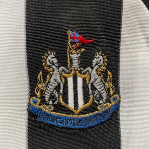 May include: Close-up of a black and white striped fabric with an embroidered crest. The crest features two seahorses, a shield with vertical stripes, and a banner that reads "Newcastle United". The crest has gold, blue, red, and silver accents.