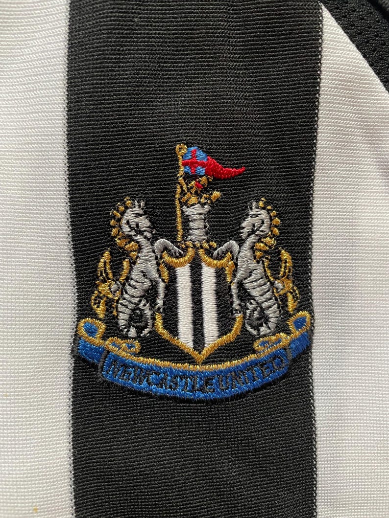 May include: Close-up of a black and white striped fabric with an embroidered crest. The crest features two seahorses, a shield with vertical stripes, and a banner that reads "Newcastle United". The crest has gold, blue, red, and silver accents.