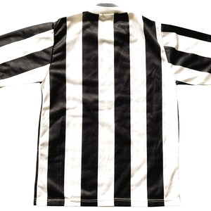 Op de afbeelding: Een zwart-wit gestreept voetbalshirt, van achteren gezien. Het shirt heeft brede verticale strepen in zwart en wit. Het heeft korte mouwen en een eenvoudige ronde hals. De stof ziet er glad uit, mogelijk een sportstof.