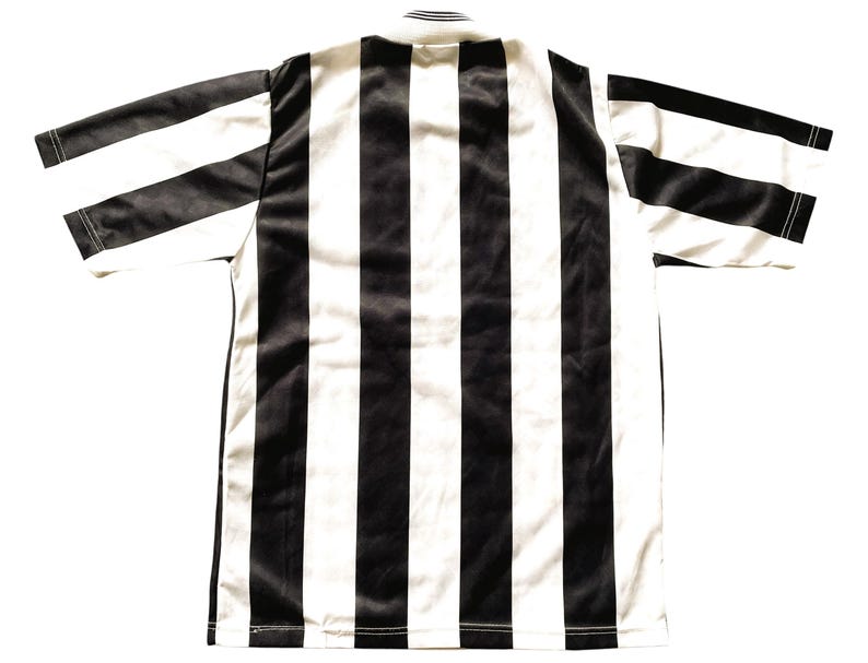 Op de afbeelding: Een zwart-wit gestreept voetbalshirt, van achteren gezien. Het shirt heeft brede verticale strepen in zwart en wit. Het heeft korte mouwen en een eenvoudige ronde hals. De stof ziet er glad uit, mogelijk een sportstof.