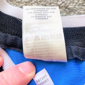 May include: A white tag with black text that reads "Die Weltmarke mit den 3 Streifen The Brand with the 3 Stripes La Marque aux 3 Bandes Obermaterial/Shell Exterieur: 100% Polyester" with washing instructions. A smaller white tag with black text that reads "687207 AVG001" is attached to a blue fabric.