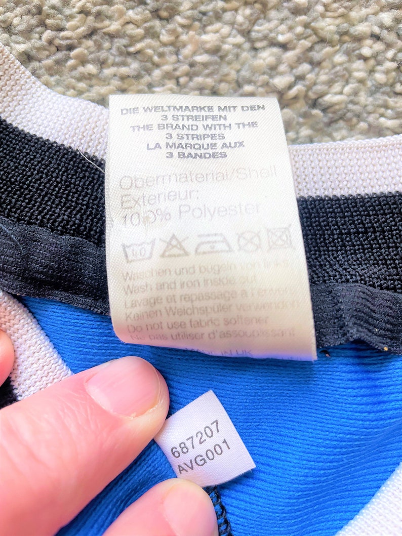 May include: A white tag with black text that reads "Die Weltmarke mit den 3 Streifen The Brand with the 3 Stripes La Marque aux 3 Bandes Obermaterial/Shell Exterieur: 100% Polyester" with washing instructions. A smaller white tag with black text that reads "687207 AVG001" is attached to a blue fabric.