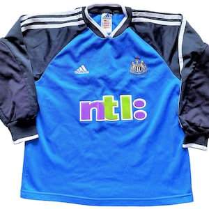 May include: A blue long-sleeved soccer jersey with black sleeves and white stripes. The jersey has the Adidas logo, the Newcastle United crest, and the NTL logo.