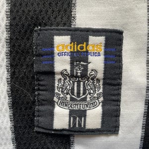 May include: Close-up of a black and white striped jersey with a Newcastle United patch. The patch features the team's crest and the Adidas logo. The jersey is a replica.