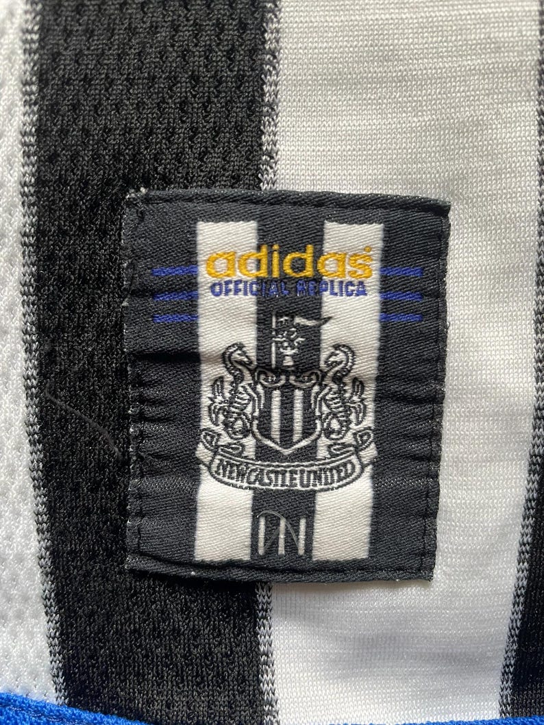 May include: Close-up of a black and white striped jersey with a Newcastle United patch. The patch features the team's crest and the Adidas logo. The jersey is a replica.