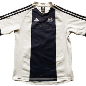 Puede incluir: Camiseta de f&uacute;tbol blanca con rayas blancas y negras y una raya vertical negra en el centro. La camiseta tiene el logotipo de Adidas y el escudo del Newcastle United en la parte delantera. El texto "nufc home" est&aacute; impreso debajo del escudo.