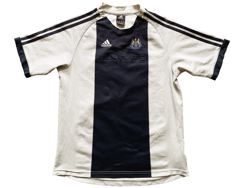 Puede incluir: Camiseta de f&uacute;tbol blanca con rayas blancas y negras y una raya vertical negra en el centro. La camiseta tiene el logotipo de Adidas y el escudo del Newcastle United en la parte delantera. El texto "nufc home" est&aacute; impreso debajo del escudo.