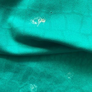 May include: Close-up of a teal fabric with visible texture and stitching. The fabric has several small, light-colored spots. The image highlights the material's weave and the color's saturation.
