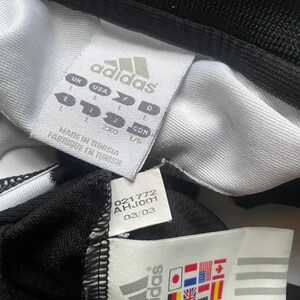 Puede incluir: Primer plano de etiquetas de ropa Adidas. Las etiquetas son blancas con texto negro y el logotipo de Adidas. Una etiqueta muestra informaci&oacute;n de tallas y la otra muestra banderas de varios pa&iacute;ses. La prenda es negra y blanca.