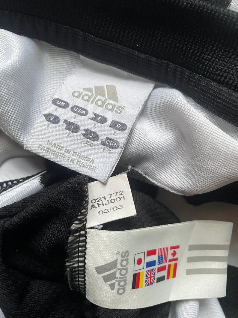 Puede incluir: Primer plano de etiquetas de ropa Adidas. Las etiquetas son blancas con texto negro y el logotipo de Adidas. Una etiqueta muestra informaci&oacute;n de tallas y la otra muestra banderas de varios pa&iacute;ses. La prenda es negra y blanca.